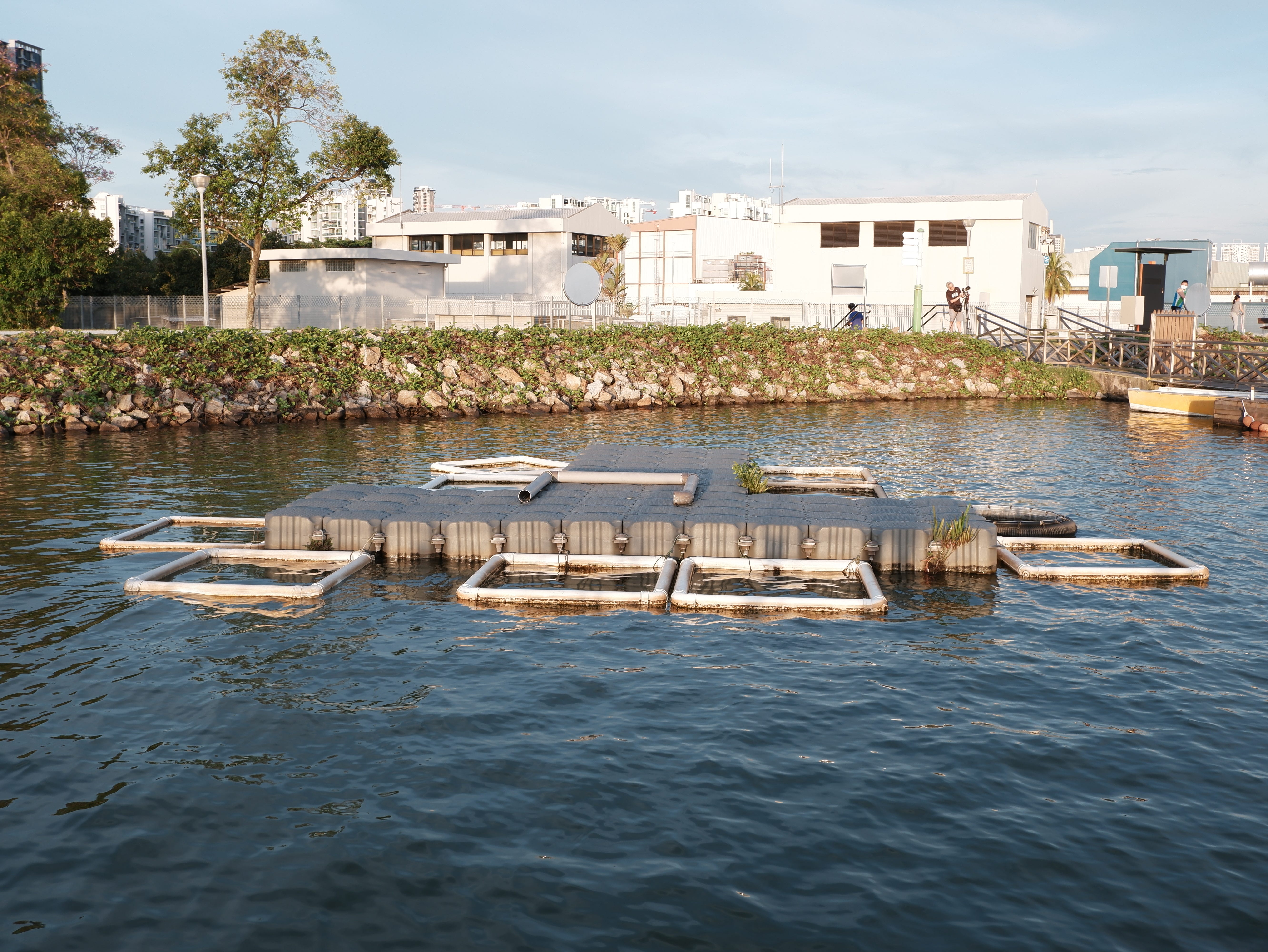 A mysterious structure floats on the water. In the distance, a photographer adjusts his tripod to capture the setting sun.