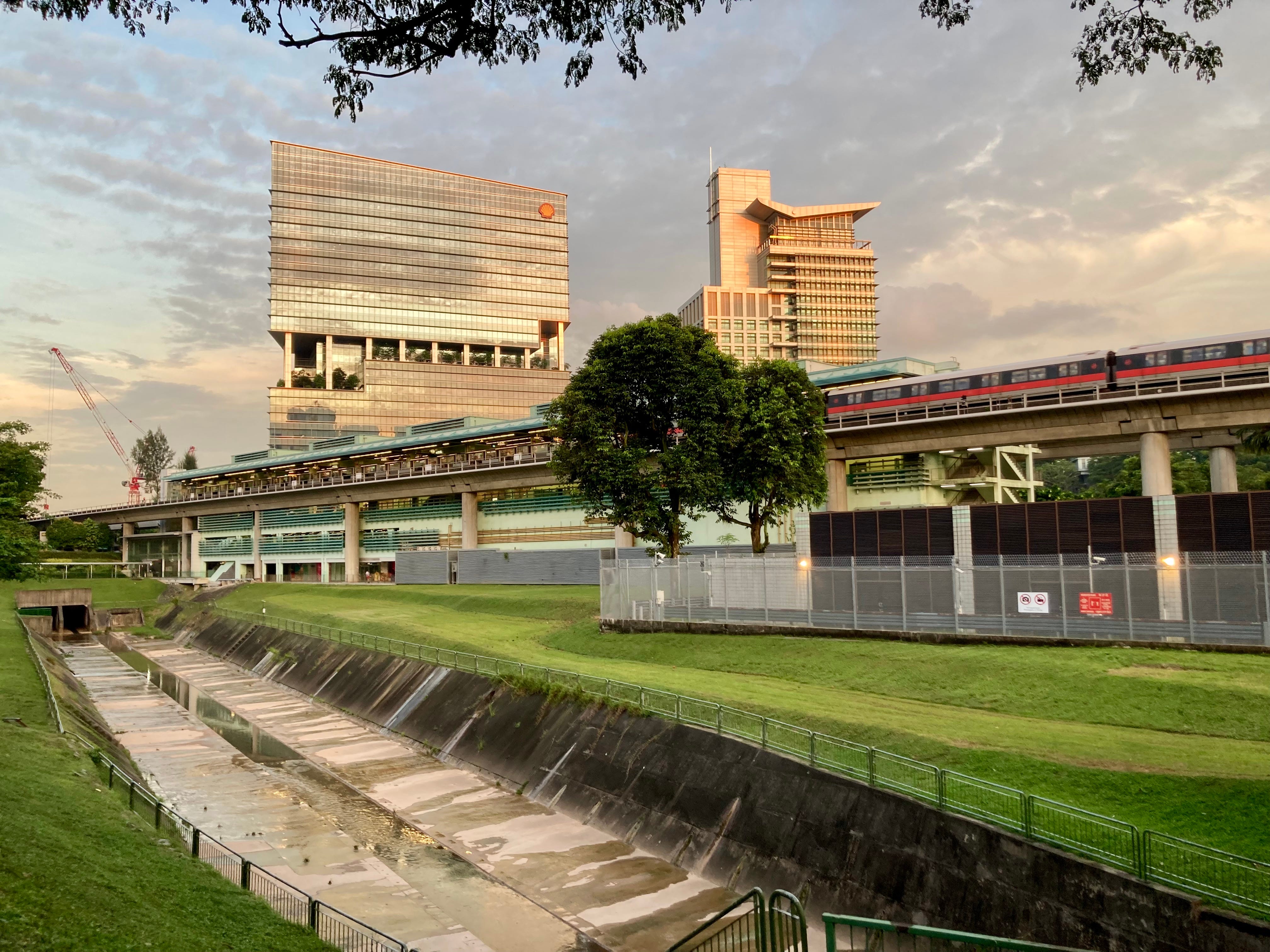 The canal emerges from a dark tunnel underneath the Buona Vista MRT station. Behind the MRT station, the Metropolis tower and the Ministry of Education office buildings catch the light from the sunset.